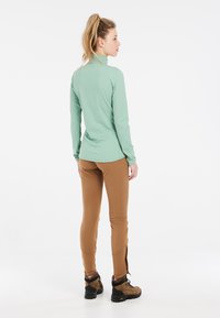 Maglia a maniche lunghe verde chiaro con colletto alto, abbinata a pantaloni aderenti marroni e scarponi da trekking marroni, vista posteriore.