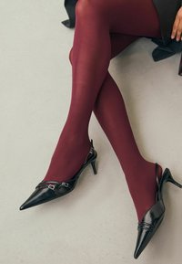 Schwarze, spitze Slingback-Pumps mit Schnallenakzenten, kombiniert mit burgunderfarbenen, blickdichten Strümpfen. Glatte Texturen und glänzende Oberfläche.