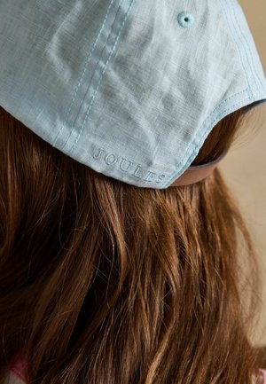 Hellblaue Stoffkappe mit gesticktem Belüftungsloch und "JOULES"-Logo, getragen auf langem braunem Haar.