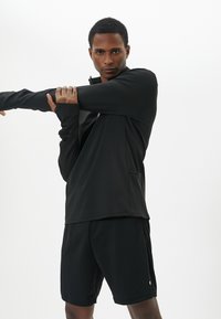 Pullover atletico nero con colletto alto, maniche lunghe e cerniera. Indossato sopra pantaloni corti neri abbinati, presenta un tessuto liscio e un design minimalista.