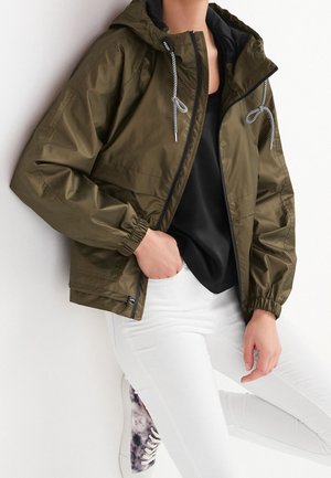 Personne portant une veste à capuche vert olive avec des cordons noirs, une chemise noire, un pantalon blanc et des baskets tie-dye violettes, appuyée contre un mur blanc.