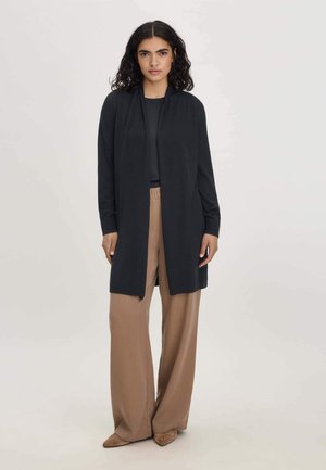 Frau mit schulterlangem lockigem Haar, die einen langen schwarzen Cardigan, ein schwarzes Oberteil, weite beige Hose und spitze beige Schuhe trägt und vor einem einfarbigen Hintergrund steht.