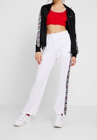 Veste noire zippée, top court rouge et pantalon de survêtement blanc avec bande logo sur le côté. Des baskets blanches complètent ce look sportif.