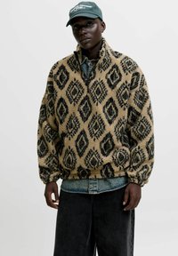 Fleece-Pullover in Beige mit schwarzem Rautenmuster, ausgestattet mit einem Reißverschlusskragen und elastischen Bündchen. Getragen über einem Jeanshemd und schwarzen Hosen.