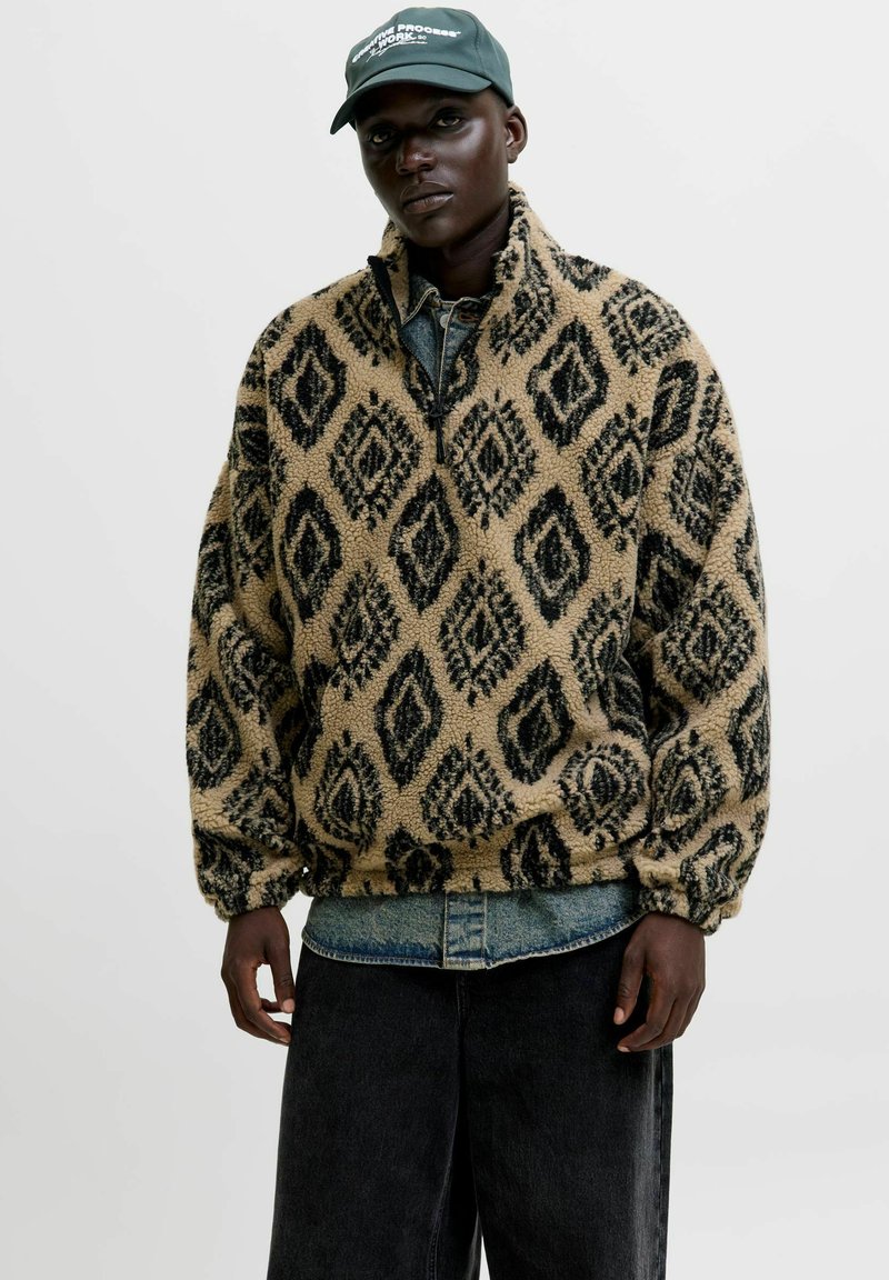 Fleece-Pullover in Beige mit schwarzem Rautenmuster, ausgestattet mit einem Reißverschlusskragen und elastischen Bündchen. Getragen über einem Jeanshemd und schwarzen Hosen.