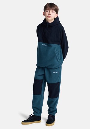 Sudadera de forro polar con la parte superior negra y la parte inferior en color teal, combinada con pantalones teal a juego con parches azul oscuro y puños elásticos.