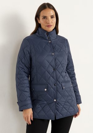Veste matelassée marine avec un col montant, fermeture à boutons-pression, deux poches avant et un motif en diamant texturé. Portée sur un pantalon noir.