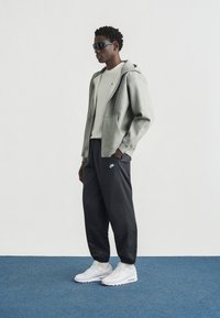 Γκρι φούτερ με φερμουάρ, μαύρο στενό παντελόνι με λογότυπο και λευκά αθλητικά παπούτσια. Λείο ύφασμα, ρυθμιζόμενη κουκούλα και πλαϊνές τσέπες.