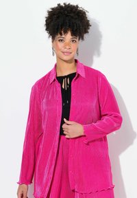 Camicia in velluto a coste rosa con colletto, maniche lunghe e chiusura con bottoni. Presenta una texture a coste e polsini arricciati, indossata sopra un top nero.
