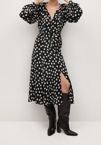 Vestido negro ajustado con un patrón geométrico blanco, mangas largas abullonadas, una abertura en la parte delantera y combinado con botas de tacón negras hasta la rodilla.