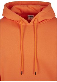 Sudadera naranja con una textura suave, que presenta una capucha con cordones, cuello ribeteado y una etiqueta de marca visible. Acabado de tejido suave.