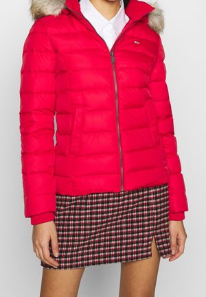 Kobieta w czerwonej kurtce puchowej z kapturem obszytym futrem, białej koszuli i krótkiej czarno-czerwono-biało kraciastej spódnicy z przednim rozcięciem.