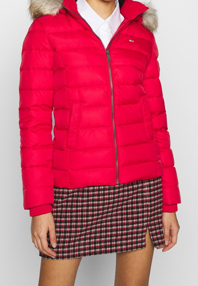 Femme portant une doudoune rouge avec une capuche bordée de fourrure, une chemise blanche et une jupe courte à carreaux noirs, rouges et blancs avec une fente à l'avant.