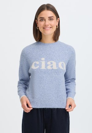 Femme aux cheveux courts bruns et boucles d'oreilles dorées porte un pull duveteux bleu clair avec des lettres blanches "ciao" et un pantalon sombre sur fond blanc.