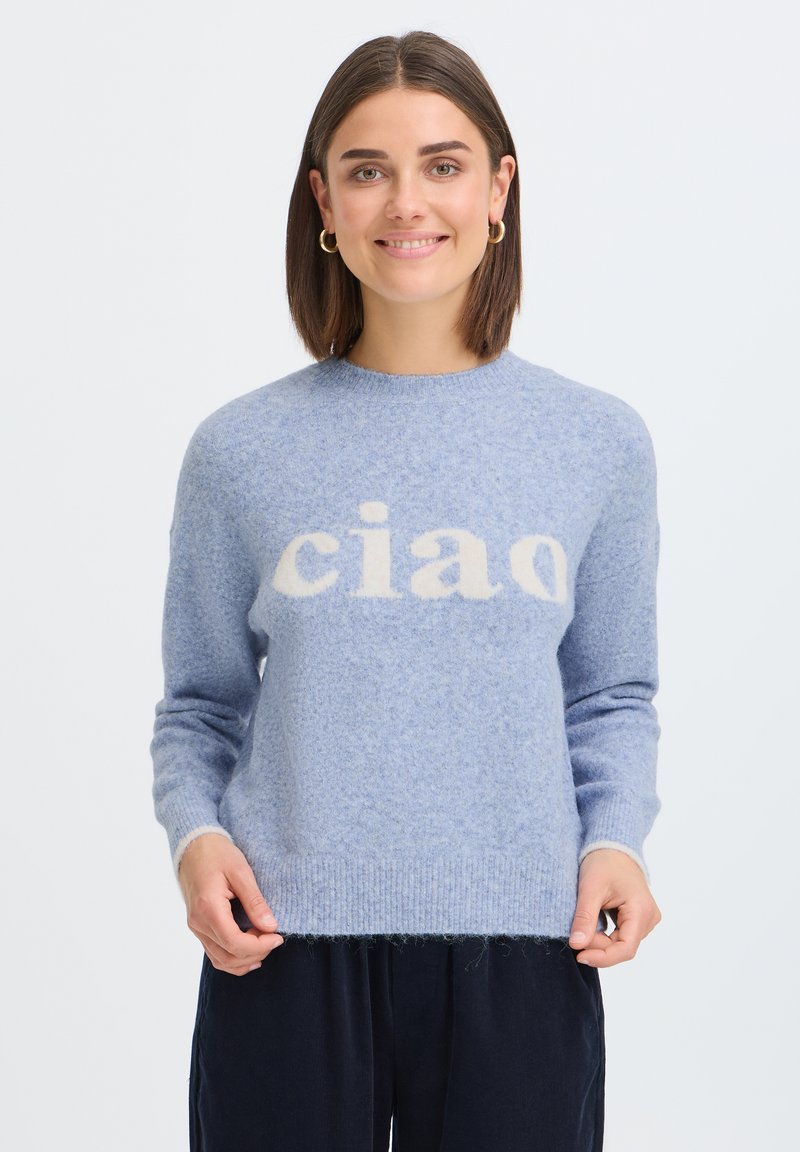 Femme aux cheveux courts bruns et boucles d'oreilles dorées porte un pull duveteux bleu clair avec des lettres blanches "ciao" et un pantalon sombre sur fond blanc.