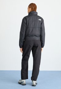 Giacca a vento nera isolata con colletto alto, design trapuntato e logo sulla schiena, abbinata a pantaloni neri e scarpe da trekking grigie.