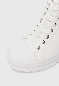 Pantof sport alb cu talpă înaltă, având o pană laterală texturată, vârf neted și șireturi albe prinse în ochiuri metalice.