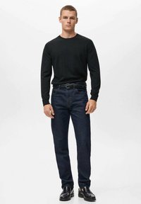 Črna majica z dolgimi rokavi, prilegajoče se jeans hlače temno modre barve, teksturiran pas in črne čevlje. Model stoji pokončno pred belo ozadje.