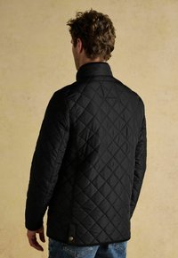 Czarna pikowana kurtka z wysokim kołnierzem i ukośnym przeszyciem, z wbudowanym zapięciem na zatrzask u dołu. Teksturowany materiał zapewniający ciepło.