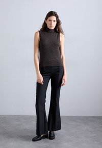 Bruine mouwloze coltrui gecombineerd met zwarte wijde broek met accentknopen; gedragen met zwarte instappers op een neutrale achtergrond.