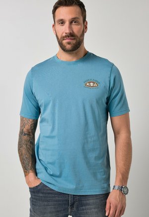 Mand med skæg iført turkis T-shirt med teksten "Hokkaido Vulkanisk Japan" og tatovering på venstre arm, står med hænderne i lommerne.