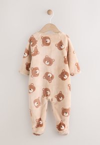 Pyjama bébé beige en polaire avec manches longues et pieds, orné d'un motif de visages d'ours marron clair et foncé, suspendu à un cintre en bois.