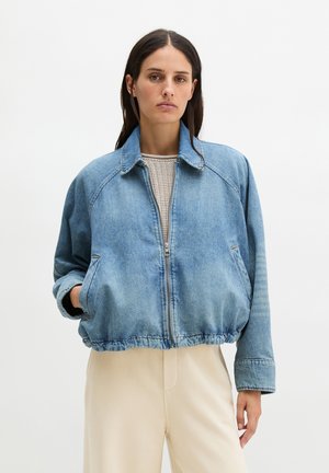 Donna che indossa una giacca di jeans azzurro con zip sopra pantaloni beige, in piedi contro uno sfondo bianco semplice con una mano in tasca.