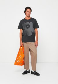 T-shirt nera con grande stampa di fiori grigi, pantaloni beige a gamba larga, tiene una borsa arancione con motivi floreali, scarpe nere, calzini bianchi.