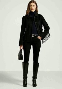 Mulher vestindo uma jaqueta preta com franjas, um suéter preto de gola alta, calças justas, botas altas pretas, segurando uma pequena bolsa preta, em pé contra um fundo simples.