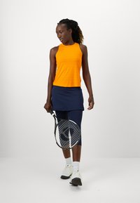 Narancssárga ujj nélküli felső, tengerészkék szoknya és leggings, teniszütővel és fehér sportcipővel párosítva. Sima textúrák, sportos fazon és réteges kialakítás.