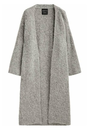 Personne portant un long cardigan en tricot gris, un top noir sans manches, un pantalon en cuir noir et des bottines noires à talons, debout de profil.