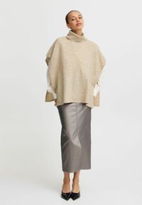 Beige gebreide oversized poncho met een hoge nek, gelaagd over een witte blouse en een metallic zilveren kokerrok, gecombineerd met zwarte hakken.
