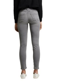 Personne portant un jean gris moulant et des baskets blanches, vue de dos sur un fond blanc.