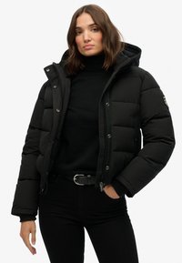 Chaqueta acolchada negra con cierre de cremallera, capucha desmontable y bolsillos. Presenta un diseño acolchado y puños de canalé para mayor calidez.