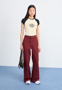 Eine junge Frau steht vor einem hellen Hintergrund, trägt ein beiges und schwarzes T-Shirt mit "Dickies"-Markenlogo, weinrote weiter Hosen und hält eine Umhängetasche.