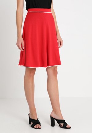 Woman wearing a knee-length red skirt with white trim and black heeled sandals, standing against a plain white background.