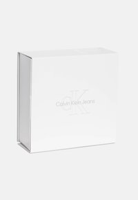 Cutie albă cu un finisaj neted, având logo-ul embosat "Calvin Klein Jeans" într-un design minimalist. Formă pătrată cu margini curate.