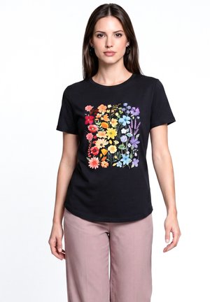 Giovane donna che indossa una maglietta nera con una varietà colorata di fiori rossi, arancioni, gialli, blu e viola stampati sulla parte anteriore.