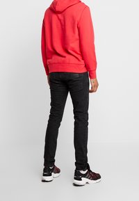 Červená mikina s kapucí, černé úzké džíny a černé sportovní boty s červenými detaily. Mikina má klokaní kapsu a žebrované manžety.