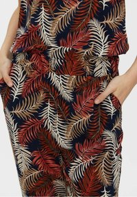 Pantalon à motif floral avec des feuilles navy, rouges et beiges. Fabriqué en tissu lisse avec des poches latérales ; taille élastique.