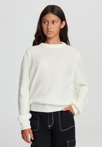 Biały sweter dzianinowy z okrągłym dekoltem, z prążkowanymi mankietami i dołem. Noszony z czarnymi spodniami z białymi przeszyciami i kieszeniami.