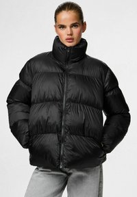 Chaqueta aislante negra con cuello alto, fabricada en un material brillante, con diseño acolchado, cremallera frontal y puños elásticos.