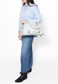 Borsa bianca texturizzata con doppio manico e tracolla, caratterizzata da molteplici tasche con zip e dettagli metallici, portata insieme a una camicia blu.
