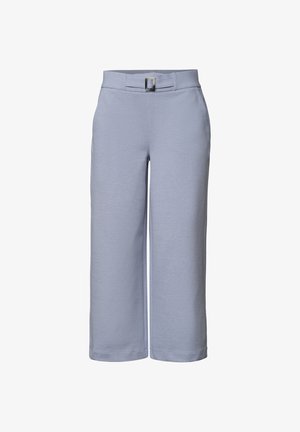 Kvinde iført lyseblå bukser med høj talje og bælte og navyblå kortærmet top, hænder afslappet ned langs siden mod en neutral baggrund.