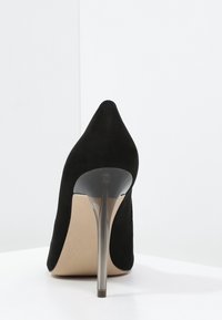 Svart mockastiletthæl sko med spiss tå, vises bakfra på en hvit overflate mot en hvit bakgrunn.