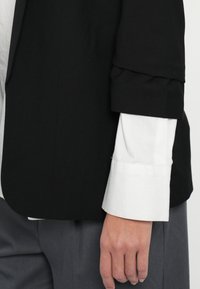 Μαύρο σακάκι με δομημένα μανίκια πάνω από λευκό πουκάμισο, με απαλή υφή και καθαρές γραμμές, συνδυασμένο με γκρι παντελόνι.