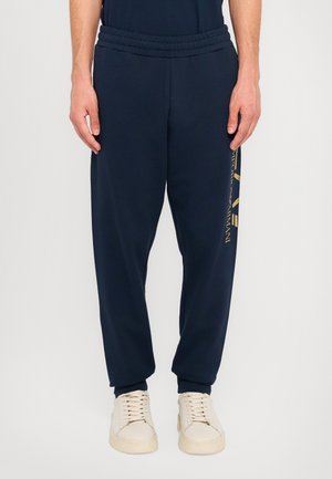 Marineblå joggebukser laget av myk stoff, med elastisk midje og en gylden logo på venstre ben.