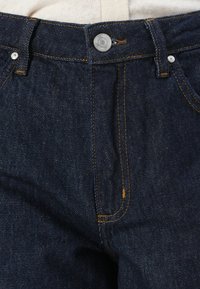 Mørkeblå denimjeans med oransje søm, med klassisk knappelukking og to frontlommer. Jeansen har en jevn tekstur; ingen synlige mønstre.