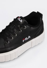 Zapatilla de cuero negra con una suela blanca gruesa, con cordones negros, ojetes plateados y un pequeño logo bordado de Fila en rojo y blanco.
