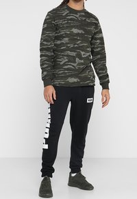 Felpa mimetica verde scuro con tessuto testurizzato e logo in rilievo, abbinata a pantaloni jogger neri con stampa bianca "PUMA" sul lato.